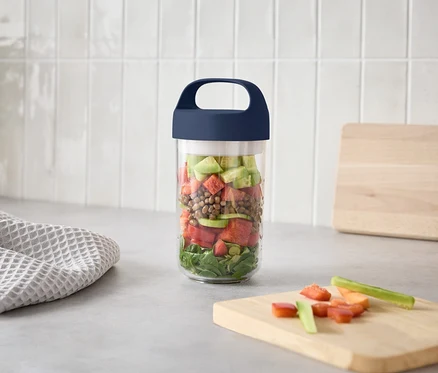Un verre avec des couches de salade est posé sur un plan de travail de cuisine à côté d'une planche à découper avec des légumes coupés.