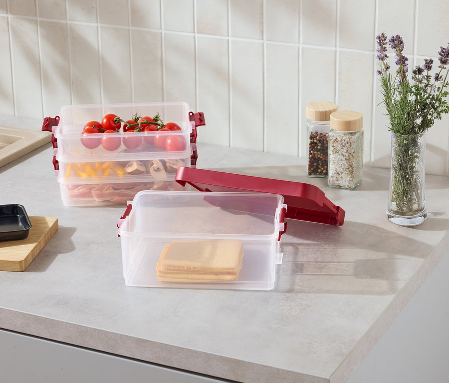 Sur un comptoir de cuisine, des récipients transparents et empilables avec des clips rouges, remplis de tranches de fromage, de pâtes, de viande et de tomates.