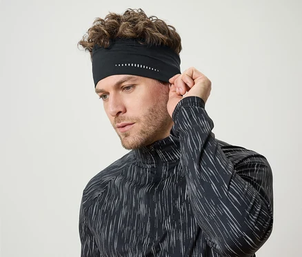 Un homme aux cheveux bouclés ajuste un bandeau de sport noir avec des détails blancs. Il porte un t-shirt noir à manches longues.