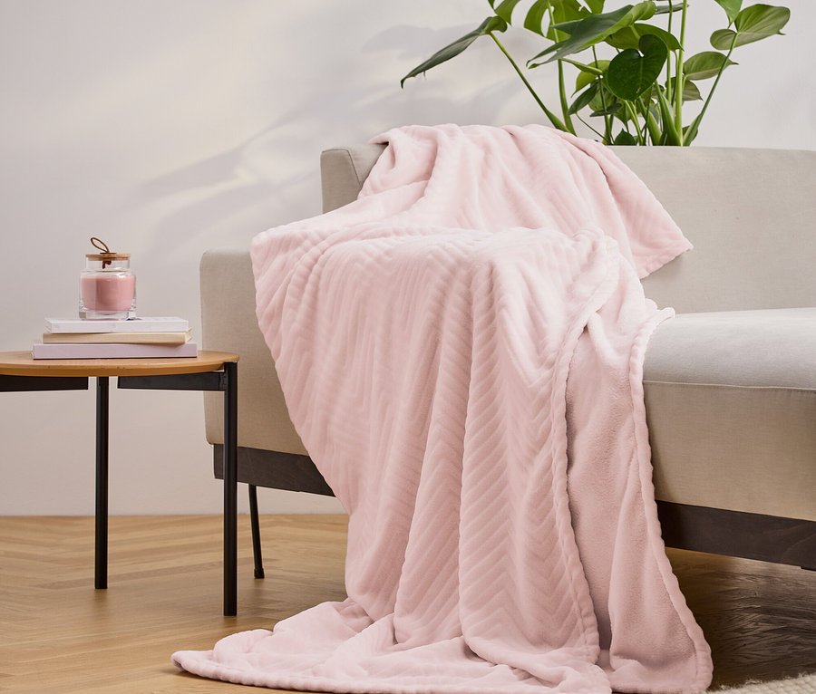 Une couverture rose texturée drapée sur un canapé beige, à côté d'une table avec des livres et une bougie, et une plante en arrière-plan.