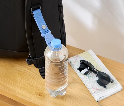 Une bouteille d'eau est accrochée à un sac à dos noir avec une bande bleue, à côté se trouvent une carte et des lunettes de soleil.