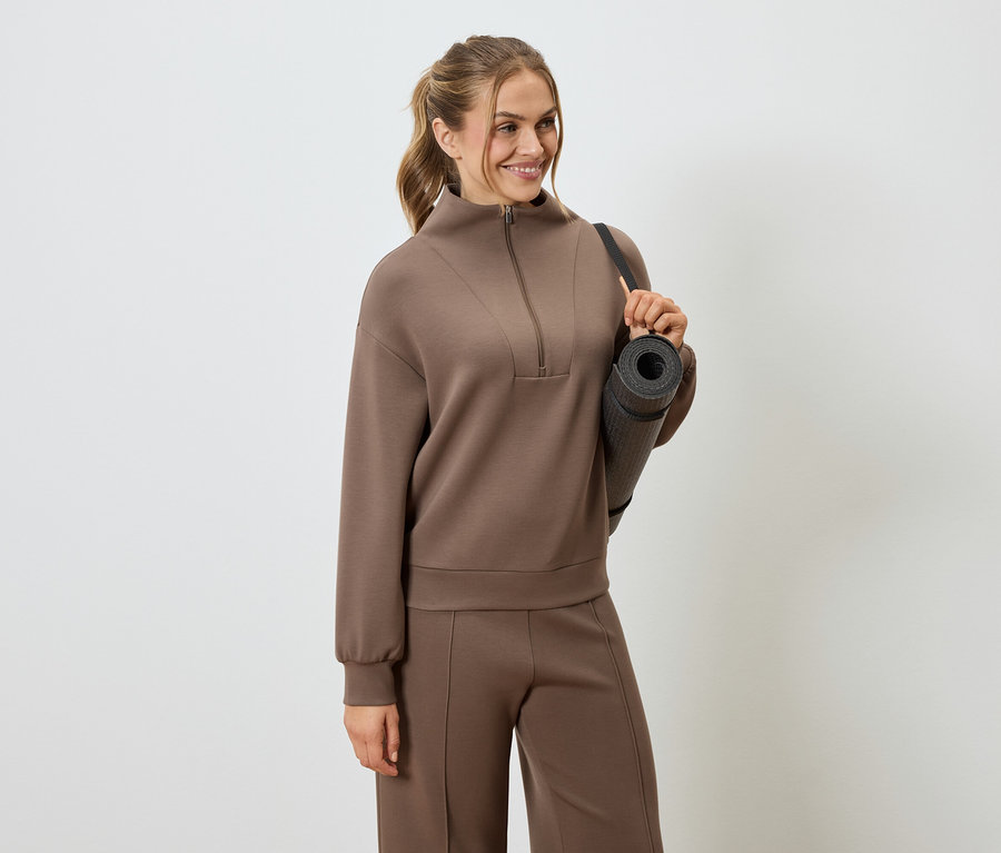 Femme portant un survêtement marron avec un tapis de yoga sur l'épaule.