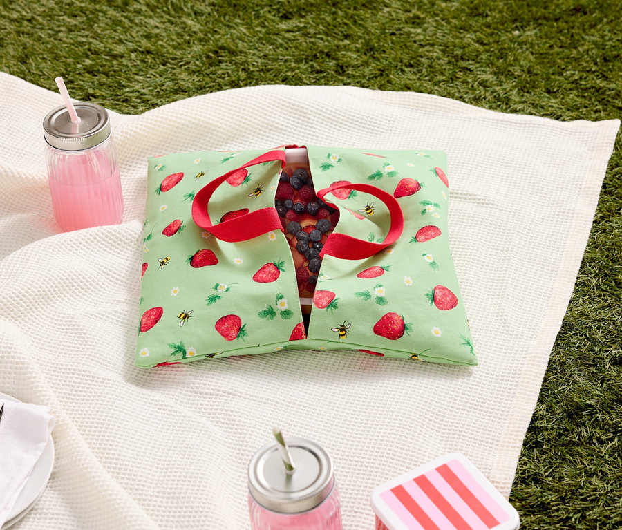 Nappe de pique-nique avec un sac à motif de fraises rempli de baies, de boissons et de couverts sur l'herbe.