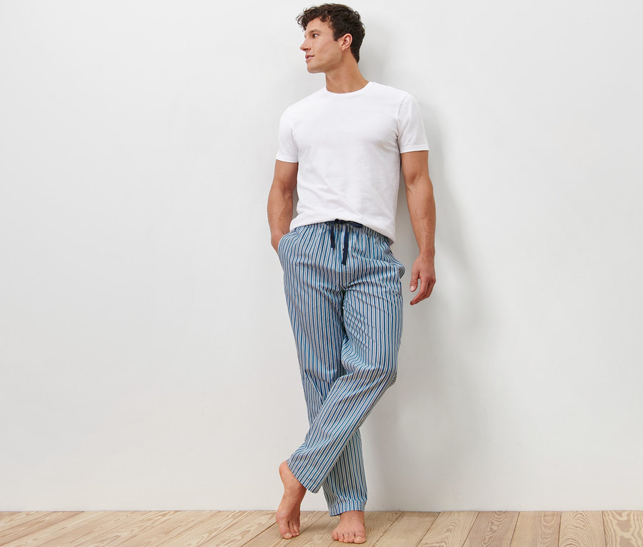 Un homme appuyé contre un mur porte un t-shirt blanc et 2 pantalons de pyjama rayés bleus.