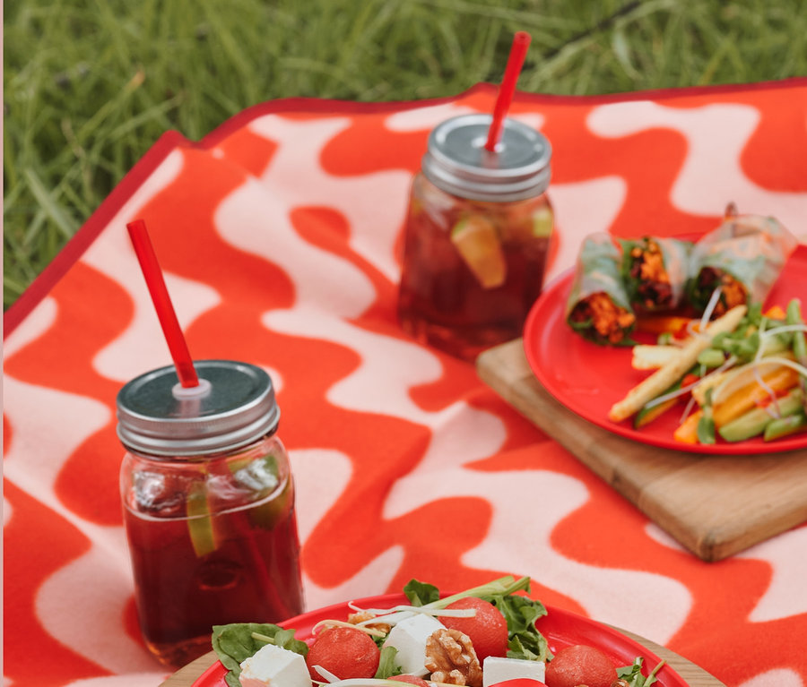 Sur une nappe de pique-nique XL sont posés une assiette de salade, des couverts de pique-nique et des verres avec des boissons et des pailles en plastique.