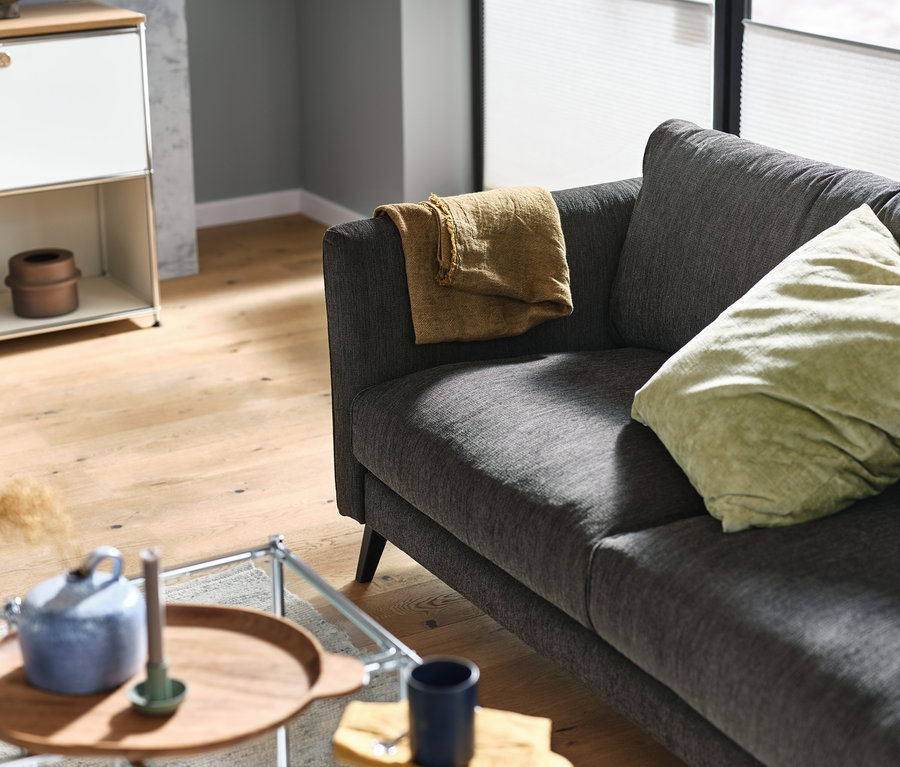 Un canapé 3 places anthracite avec une couverture et un coussin beige. Sur la gauche, une table basse à roulettes « CN3 » blanche et un buffet en métal « CN3 » blanc.