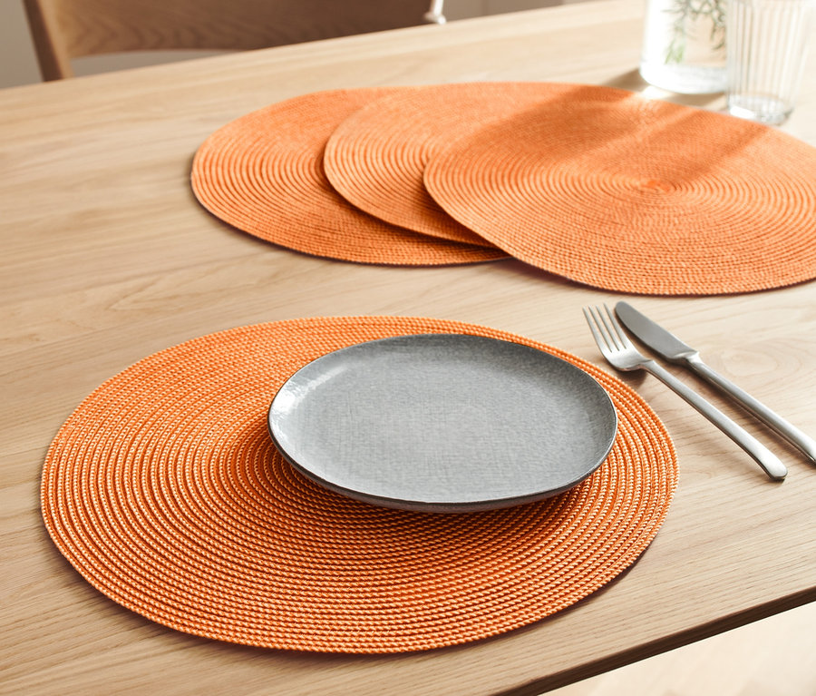 Sur une table, il y a 4 sets de table orange. Sur l'un d'eux, il y a une assiette grise, un couteau et une fourchette.