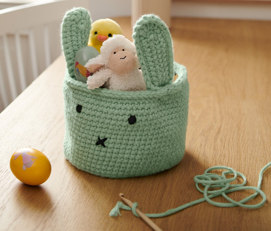 Un panier de Pâques décoratif en crochet vert menthe en forme de lapin avec un visage. À l'intérieur, il y a un poussin jaune, un mouton et un œuf peint. À côté, il y a un autre œuf peint et un crochet avec de la laine.