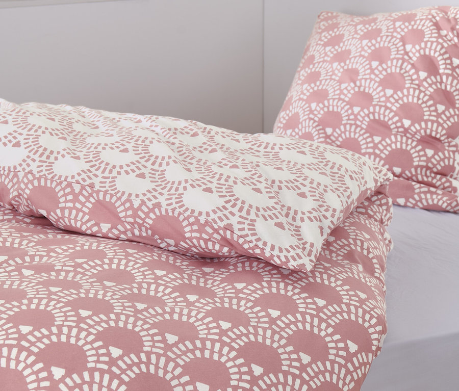 Gros plan d'une parure de lit en jersey rose avec un motif. On aperçoit deux paniers en osier et des livres en arrière-plan.