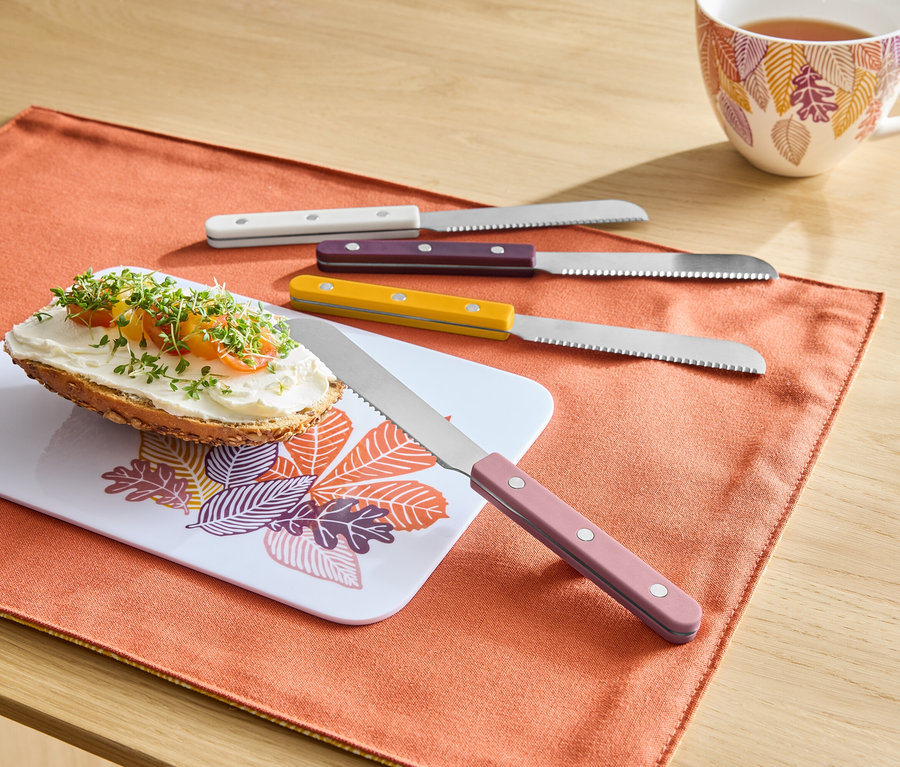 Sur un set de table orange se trouvent une tartine avec de la pâte à tartiner sur une planche à motif de feuilles, à côté se trouvent quatre couteaux avec des manches colorés et à l'arrière-plan une tasse de thé.