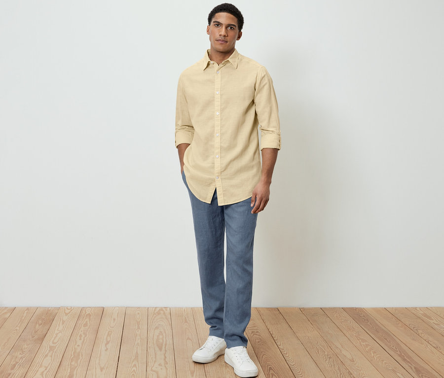 Un homme pose en chemise jaune en lin, pantalon bleu et chaussures blanches.