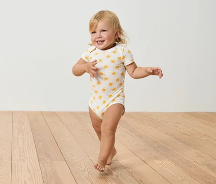 Un bébé blond avec un body à motifs de soleil marche sur un plancher en bois.