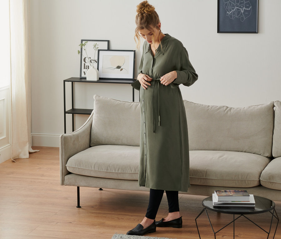 Une femme enceinte noue une robe-chemise de grossesse olive dans un salon.