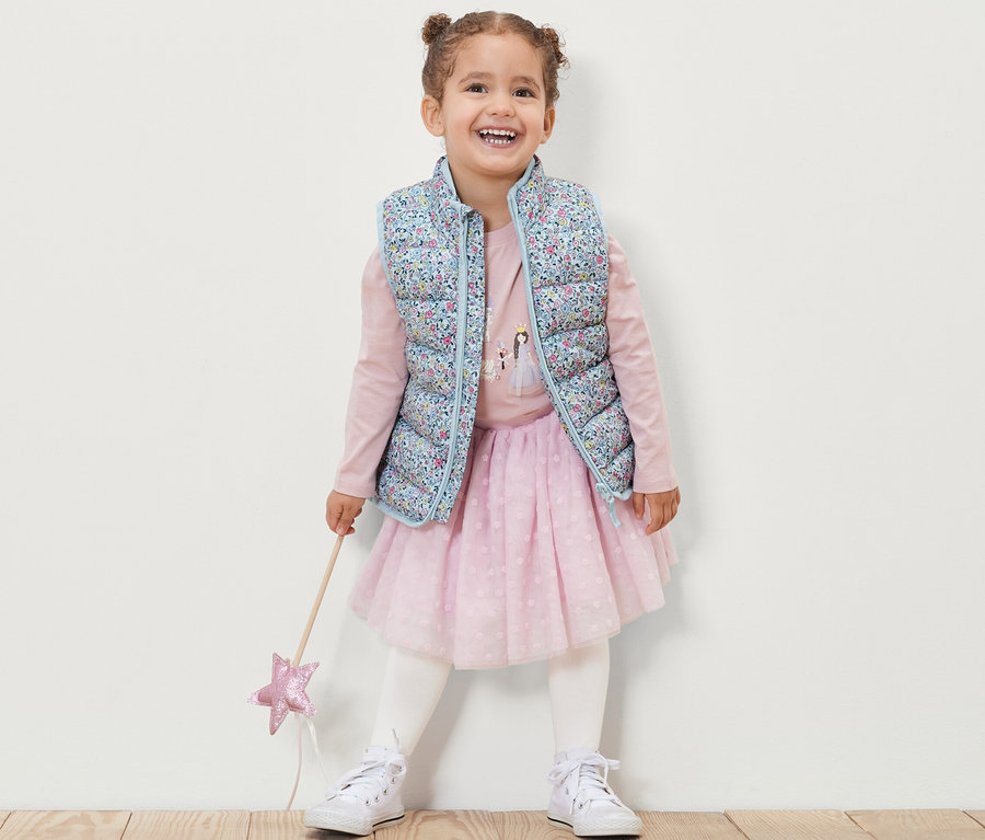 Une fillette souriante porte un gilet sans manches matelassé à fleurs, un t-shirt rose à manches longues, une jupe rose et des baskets blanches, et tient une baguette magique.