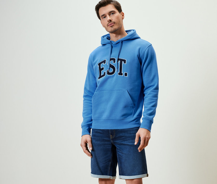 Homme posant en sweat-shirt à capuche bleu et short en jean.