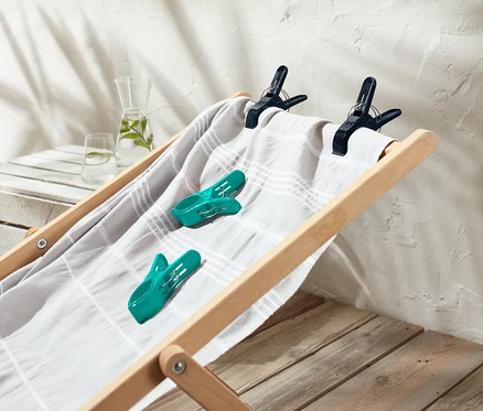 Sur un transat en bois se trouvent une serviette rayée gris et blanc, deux pinces à linge turquoise et deux noires.
