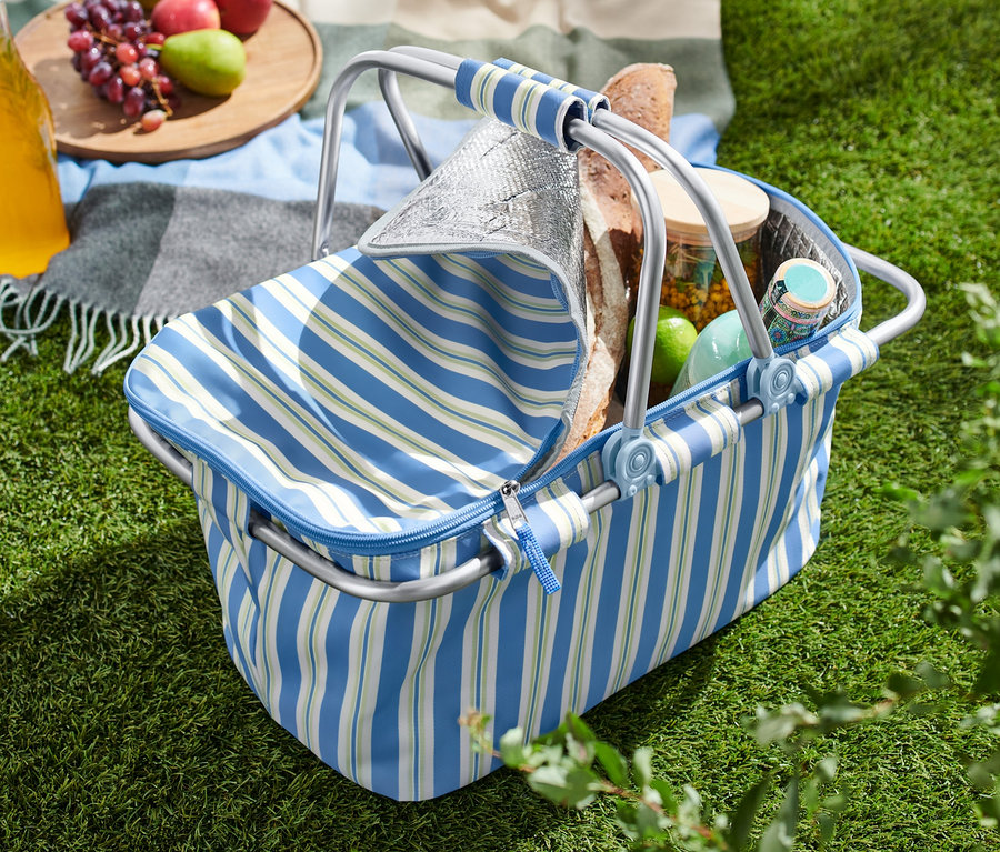 Un panier isotherme ouvert sur l'herbe à côté d'une couverture avec des pommes, des raisins et une boisson.