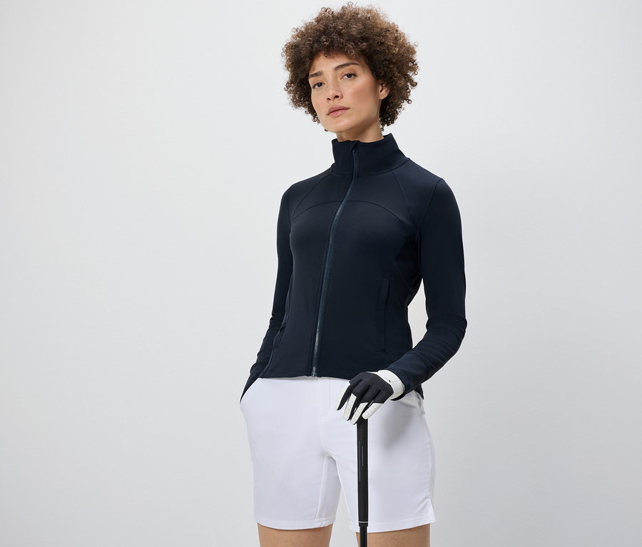 Femme portant une veste fonctionnelle, un short blanc et tenant un club de golf.