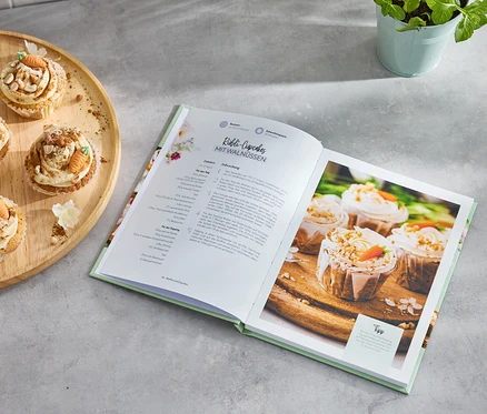 Quatre cupcakes aux carottes sur un plateau en bois à côté d'un livre de recettes ouvert intitulé «
Petit bonheur de pâtisserie
».