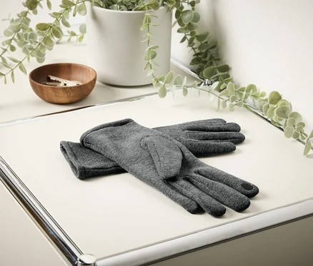 Des gants gris sont posés sur une table blanche. En arrière-plan, on aperçoit un bol en bois avec des clés et une fleur dans un pot blanc.