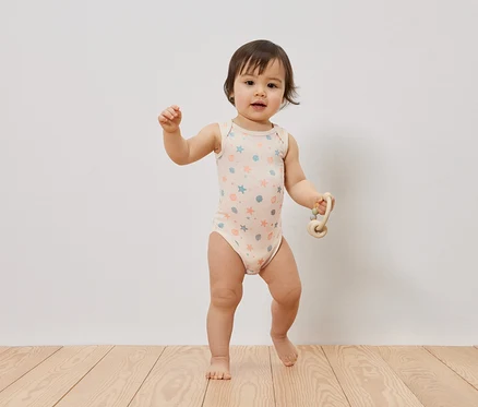 Un bébé est debout pieds nus sur un plancher en bois, portant un body bébé rose pâle sans manches avec des étoiles et des coquillages bleus et oranges, et tient un hochet clair.