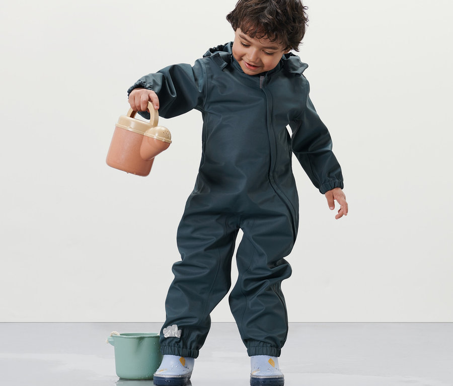 Un enfant en une-pièce imperméable pour tout petit tenant un arrosoir, debout à côté d'un seau.