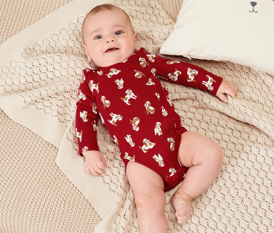 Bébé allongé dans 2 bodys pour bébé sur une couverture en tricot beige à côté d'un coussin blanc avec un visage d'ours.