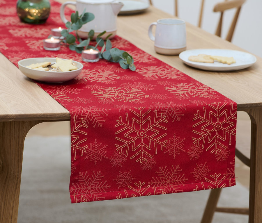 Sur une table en bois, un chemin de table en jacquard rouge à motif de flocons de neige est disposé avec des assiettes de biscuits, des bougies et une théière.