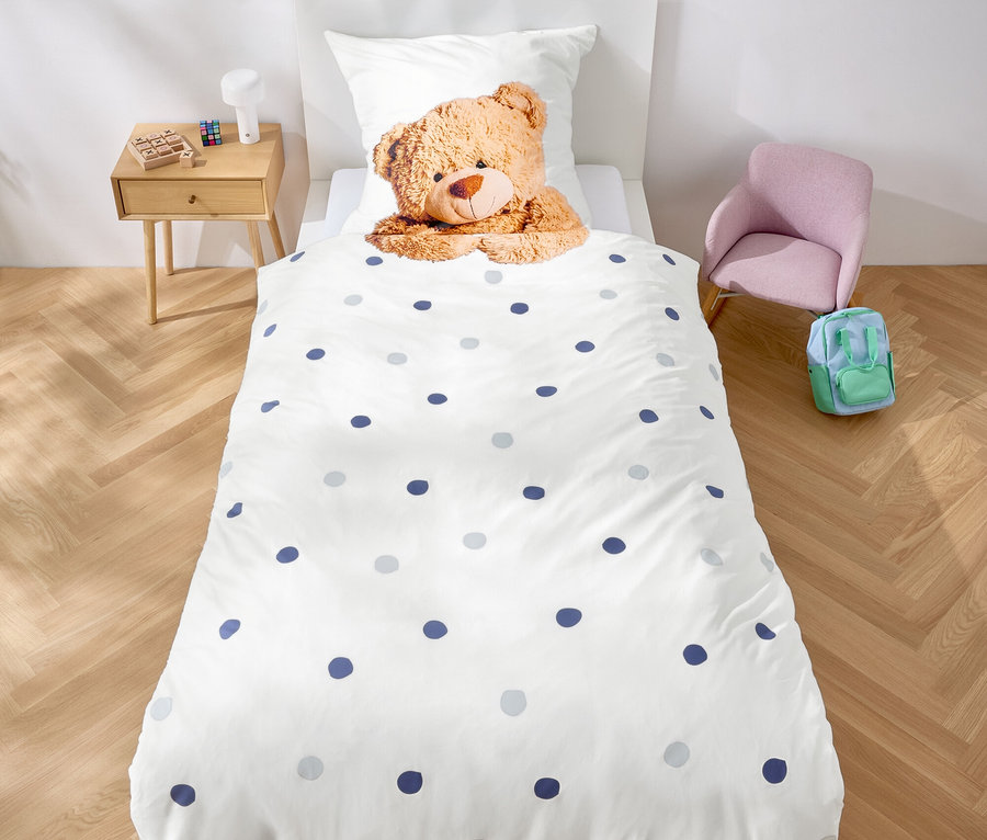 Dans la chambre, il y a un lit avec des draps blancs à pois bleus et un ours en peluche. Une guirlande de triangles violets est accrochée au-dessus du lit.