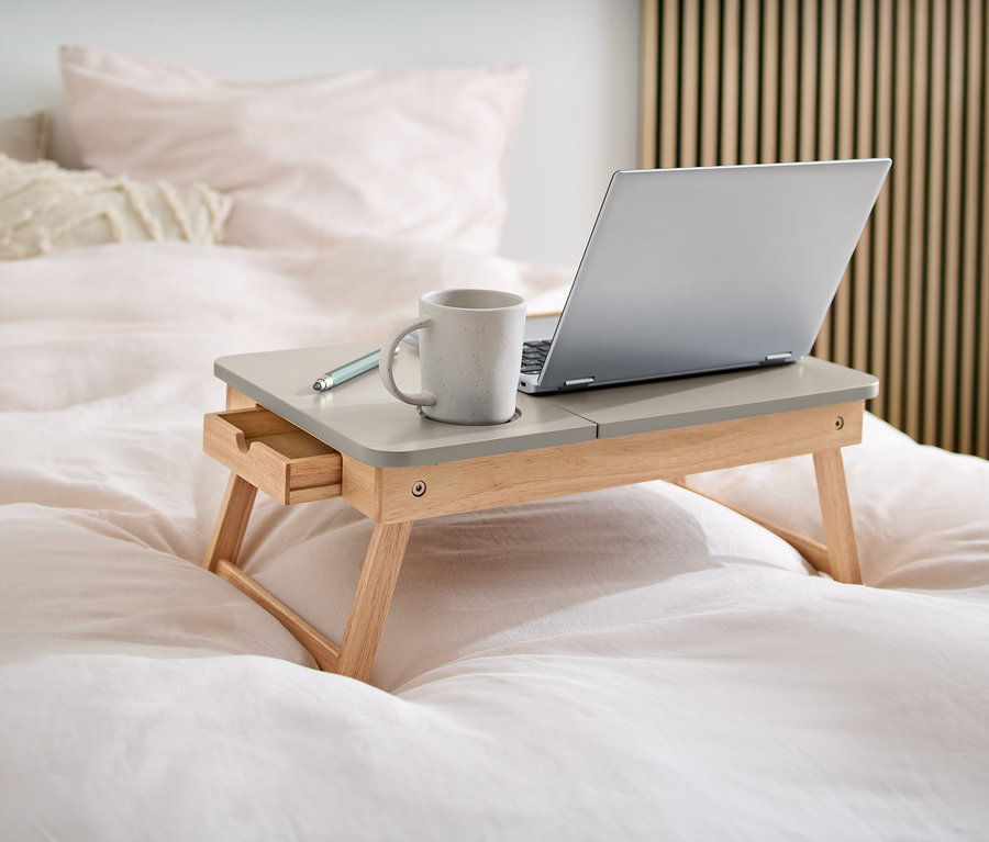 Sur un lit se trouve une table en bois pour ordinateur portable, sur laquelle sont posés un ordinateur portable, une tasse et un stylo.
