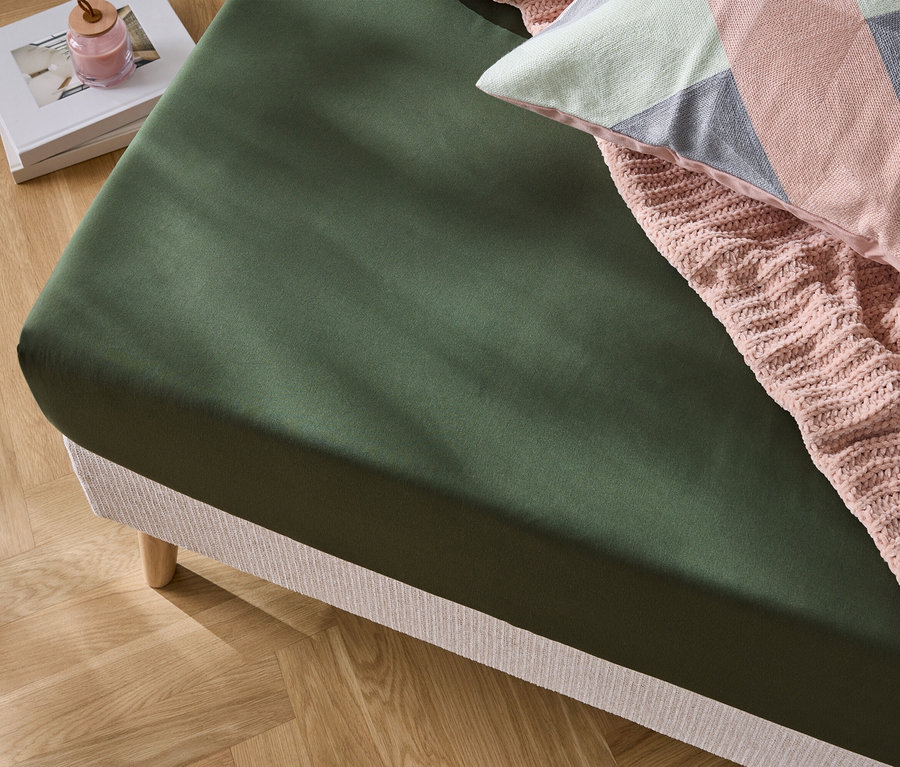 Gros plan d'un lit avec un drap-housse vert olive, une couverture en tricot rose et un oreiller décoratif.