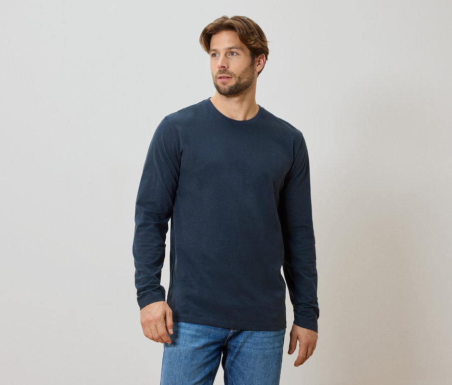 Un homme barbu porte un t-shirt bleu marine à manches longues et un jean.
