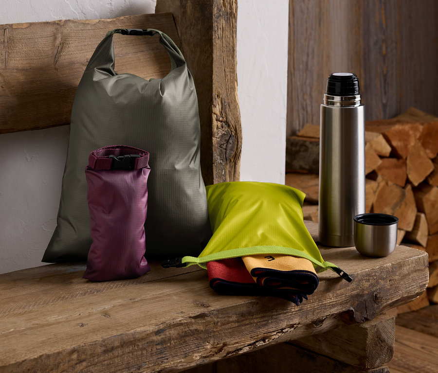 Sur un banc en bois reposent un sac vert, un sac violet, un sac vert avec des serviettes, une thermos et une pile de bois.