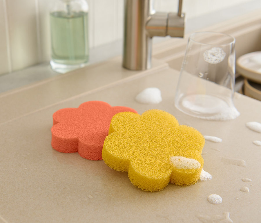 Sur l'égouttoir reposent deux éponges en silicone en forme de fleur, un verre et de la vaisselle sont recouverts de mousse.