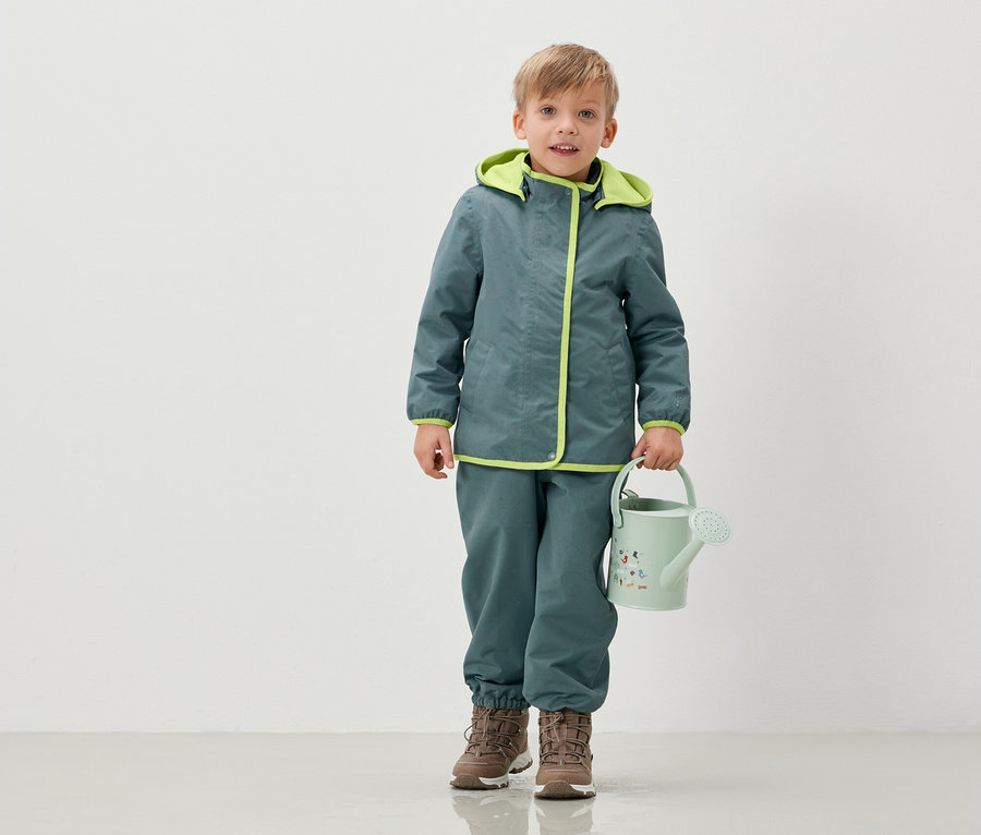 Un garçon en veste et pantalon tient un arrosoir vert clair.