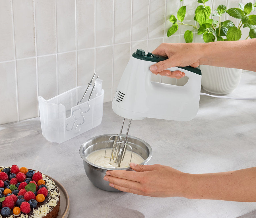 Une femme mélange de la pâte dans un bol avec un batteur à main à côté d'un gâteau aux fruits.
