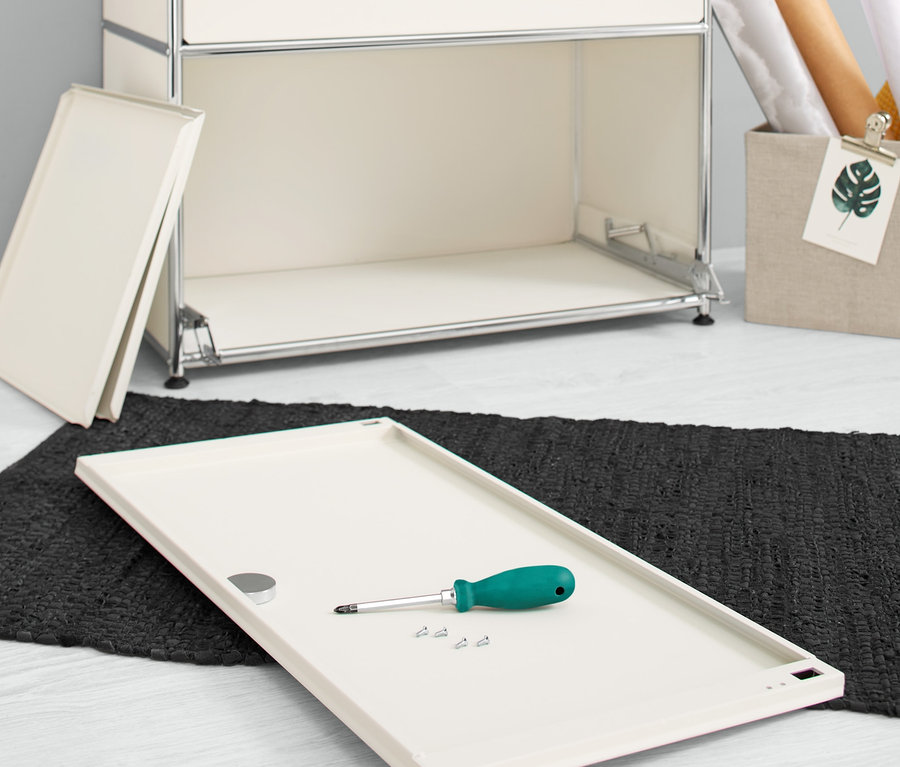 Un jeu d'abattants supplémentaires en métal « CN3 », blanc, repose sur le sol avec un tournevis et des vis à côté d'une commode.