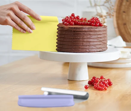 Une main utilise un grattoir jaune du lot d'ustensiles de pâtisserie pour lisser un gâteau au chocolat avec des groseilles rouges sur un socle blanc.