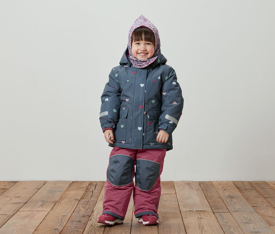 Une petite fille souriante se tient sur un plancher en bois, portant une veste d'hiver bleue à motifs de cœurs, un pantalon rose et un foulard sur la tête.