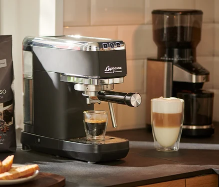 Un expresso coule d'une machine à porte-filtre « Lapressa », noire, dans un verre. Un moulin à café et un paquet de café sont visibles à l'arrière-plan.