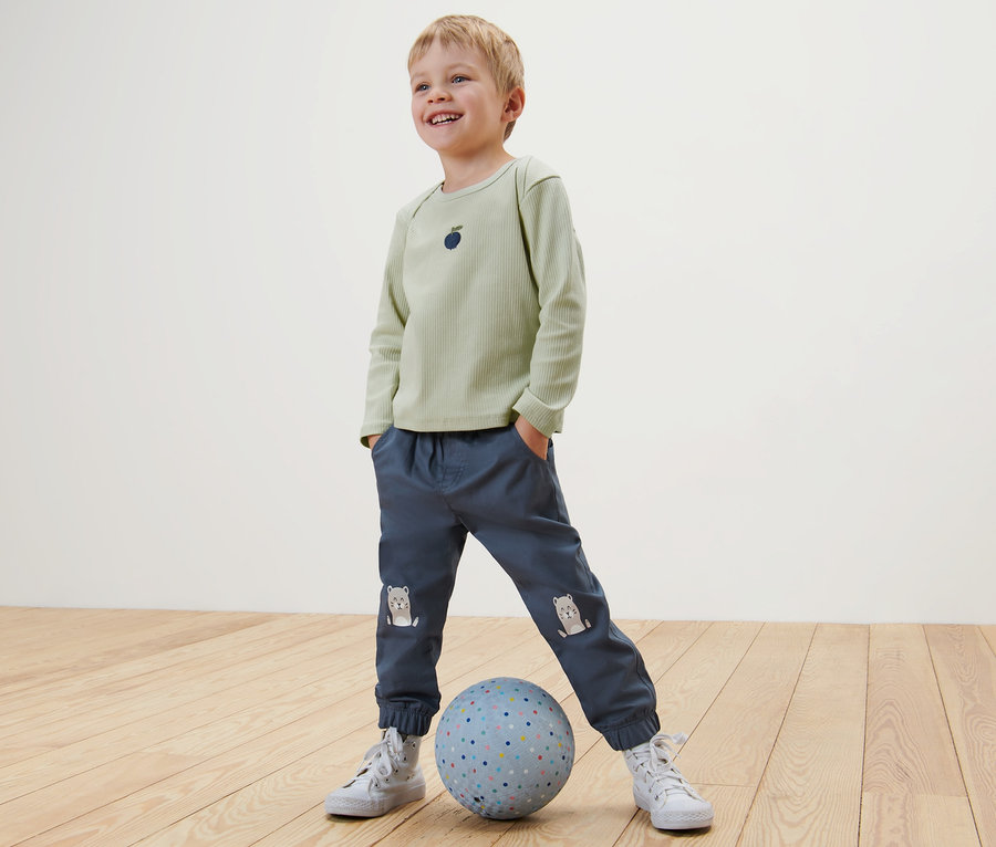 Un garçon se tient debout, les mains dans les poches, un pied sur un ballon. Il porte un t-shirt vert clair et un pantalon bleu Pantalon à enfiler pour enfant – Fit «
Ella
», loutre.