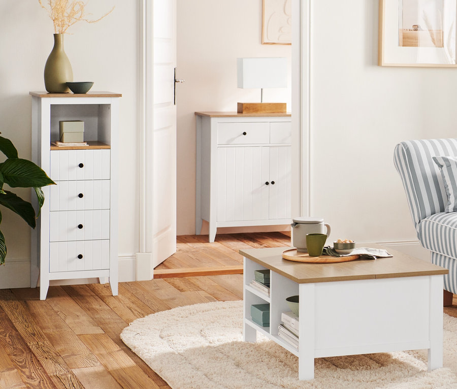 Dans le salon, un lampadaire en bois « Trépied » est placé à côté d'un canapé et d'une table basse repliable. À l'arrière-plan, on trouve une commode à tiroirs et une lampe de table avec pied en bois.