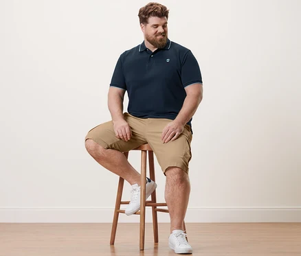 Un homme est assis sur un tabouret et porte un polo en piqué bleu marine et un short cargo sable.