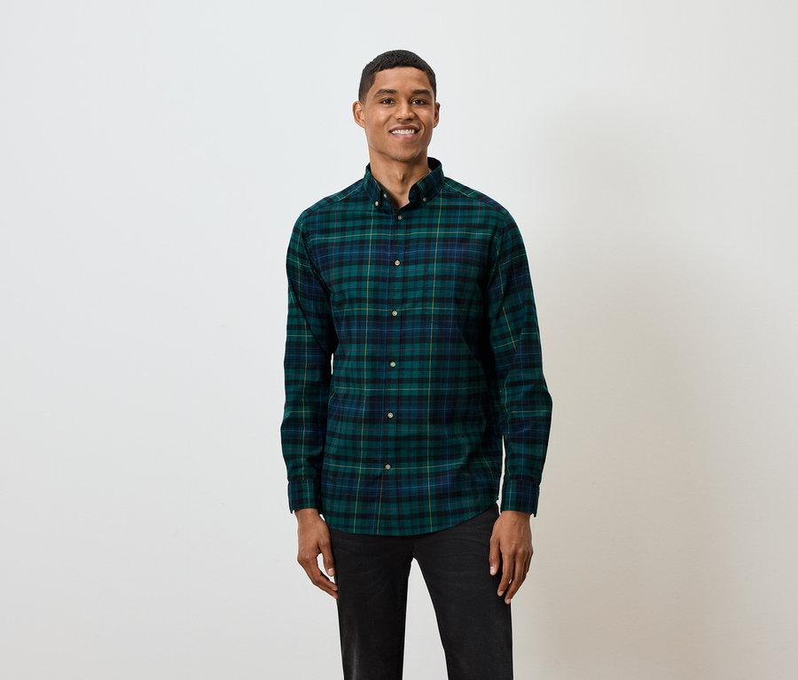 Un homme aux cheveux courts pose sur un fond blanc, portant une chemise à manches longues à carreaux verts et bleus et un jean noir.