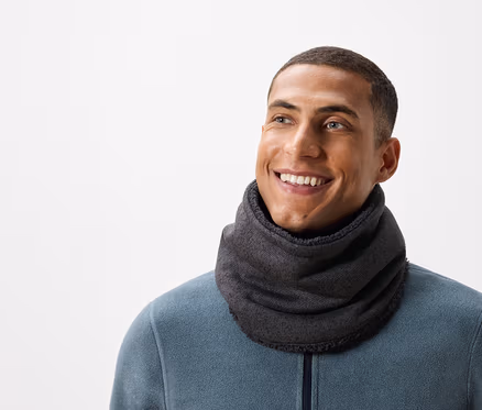 Un homme souriant aux cheveux courts porte un cache-col gris et un pull bleu-vert.