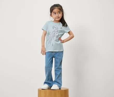 Une fille se tient sur un socle en bois. Elle porte un jean bleu et un t-shirt bleu clair avec un imprimé d'Elsa de La Reine des neiges.