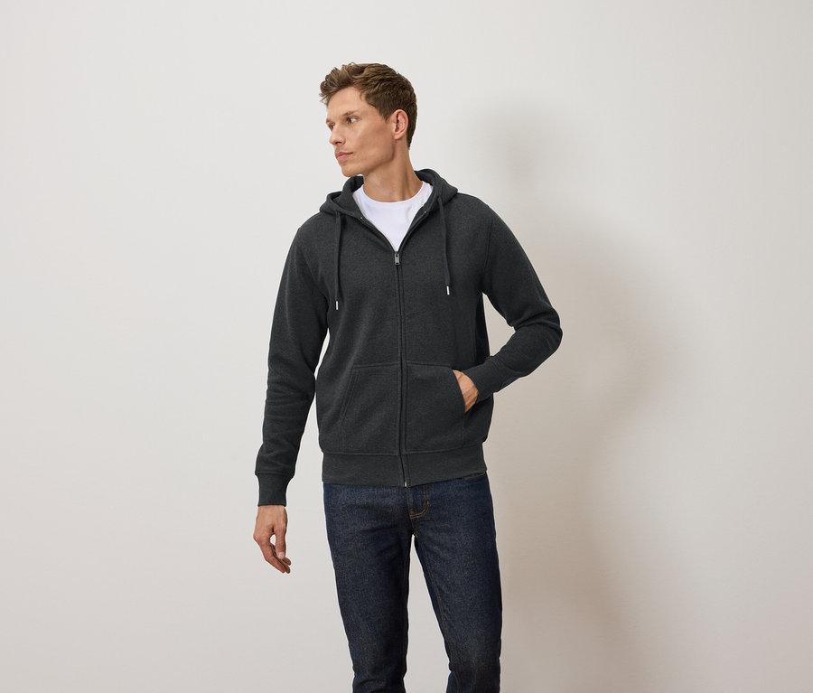 Un homme porte un sweat à capuche zippé gris foncé, un t-shirt blanc et un jean bleu.