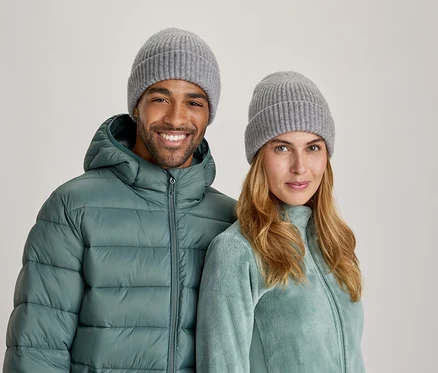 Un homme et une femme souriants portent des bonnets tricotés gris. L'homme porte une doudoune turquoise et la femme une veste polaire turquoise clair.