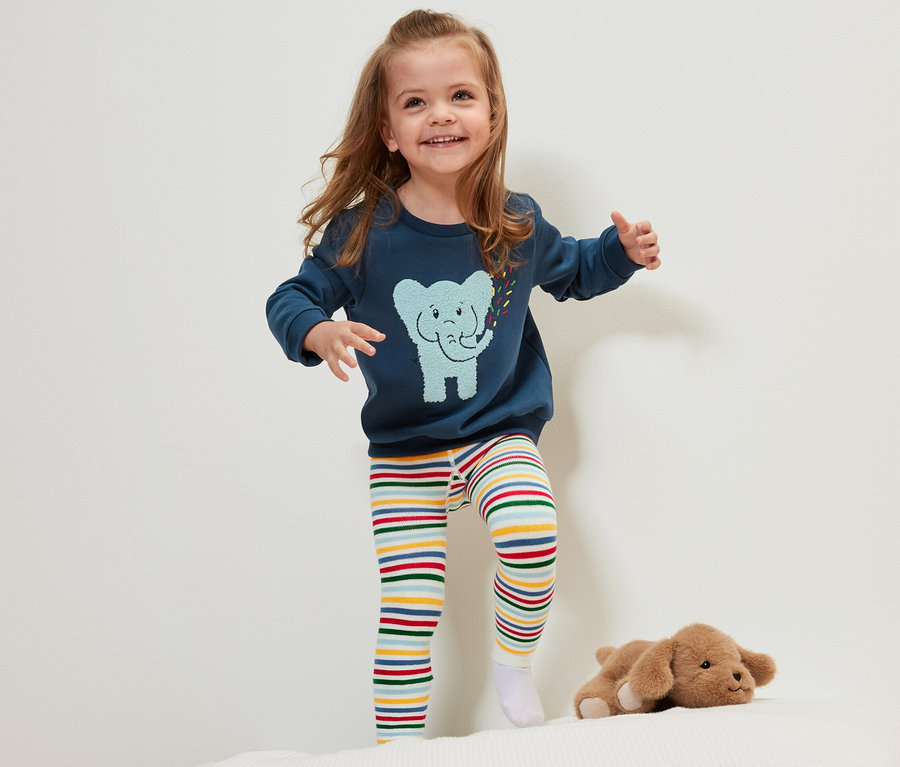 Une petite fille souriante se tient sur un lit et porte l'un des 3 leggings thermiques pour enfant à rayures colorées.