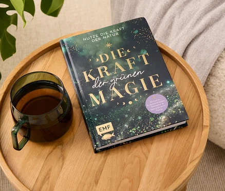 Sur un plateau en bois reposent un livre intitulé "Die Kraft der grünen Magie" et une tasse en verre contenant une boisson.